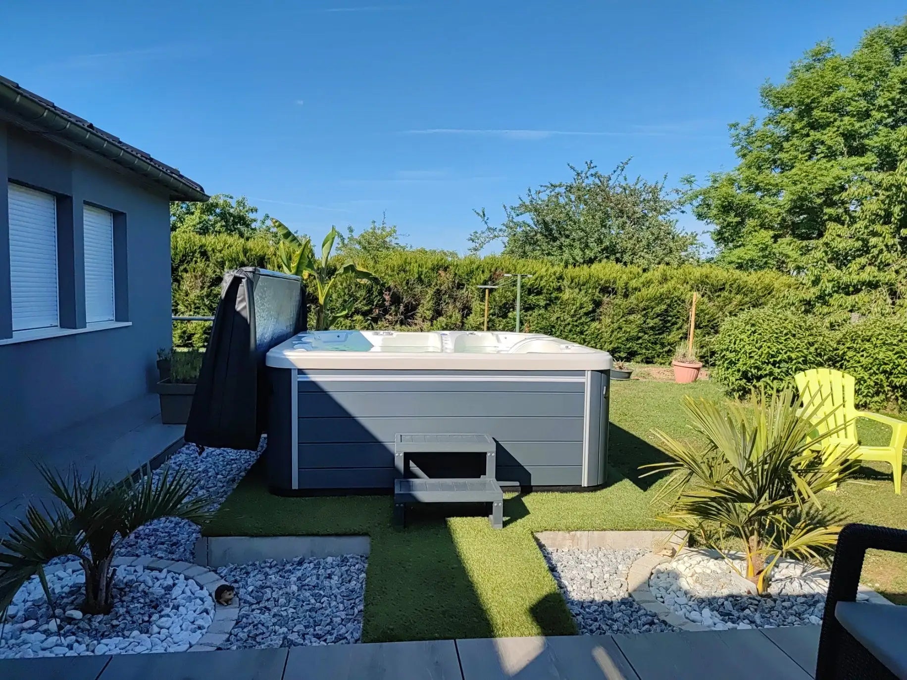 Bain à remous rectangulaire gris avec un dessus blanc et un couvercle noir partiellement ouvert, situé sur de l'herbe artificielle entourée de cailloux et de verdure.